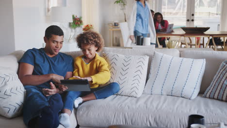 mum joins husband and daughter playing with tablet in living room, grandparents in the background