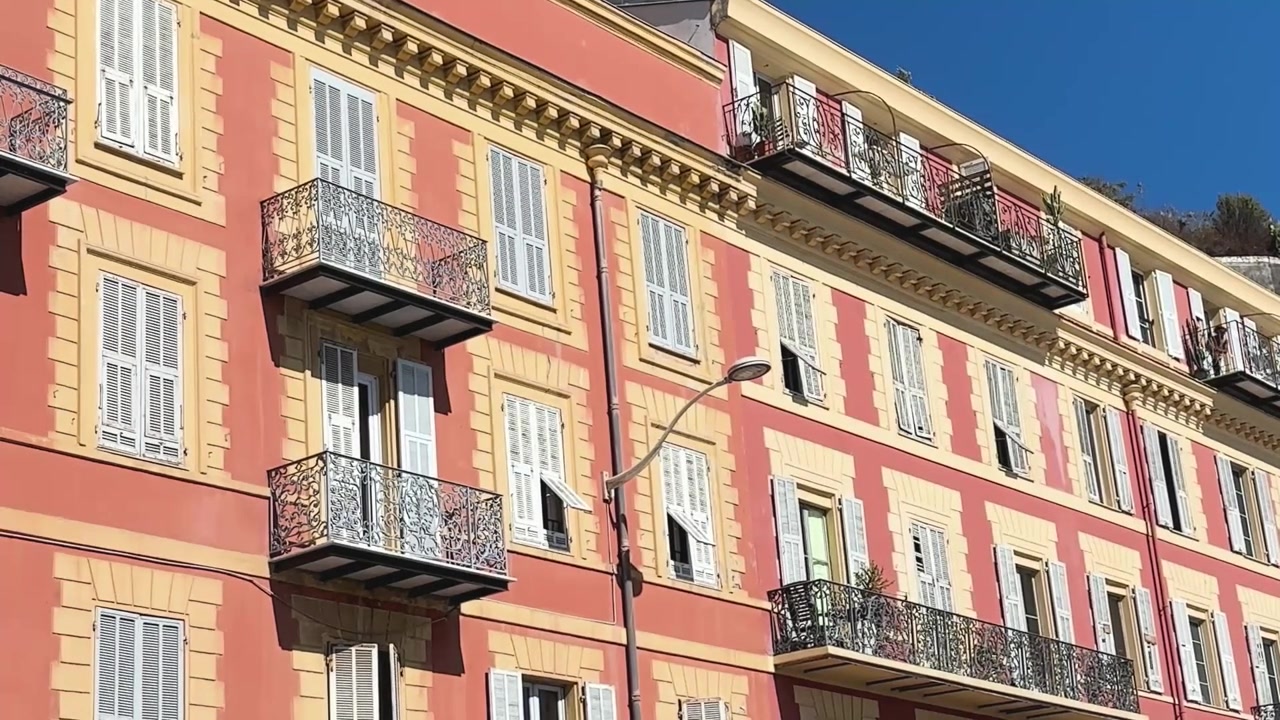 Facciata residenziale color corallo e giallo illuminata dal sole con persiane bianche e balconi in ferro battuto ornati a Nizza, Francia, sulla Costa Azzurra