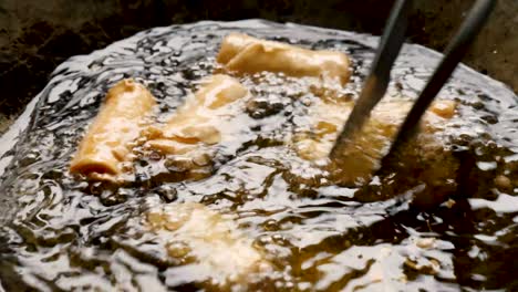 Homemade-fried-spring-rolls-frying,-cooking-and-turned-in-hot-boiling-oil-with-metal-tongs,-static-close-up