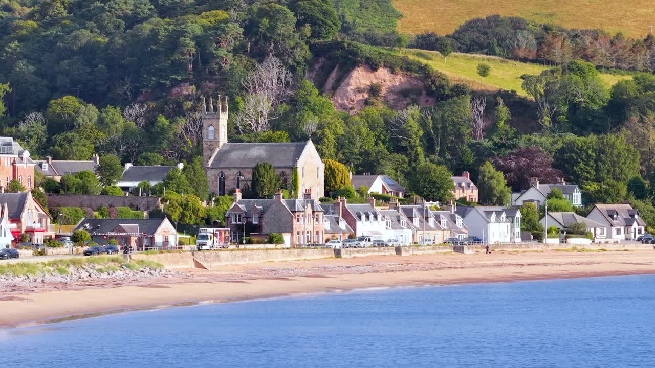 Slow lateral pan across Rosemarkie’s coastal village, historic church, green hills, and sandy shoreline