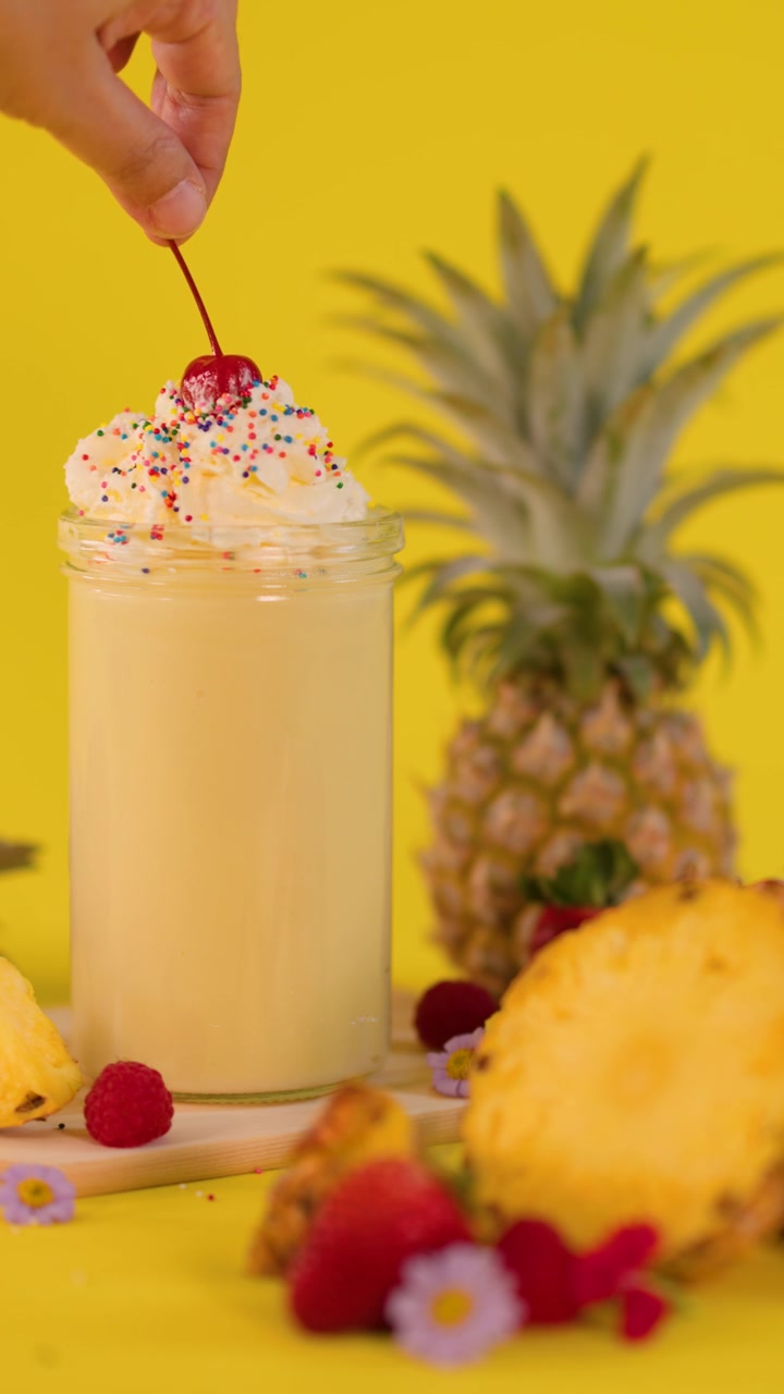Una mano posiziona una ciliegia su un milkshake all'ananas con panna montata, circondato da frutta tropicale, sotto un'illuminazione brillante.