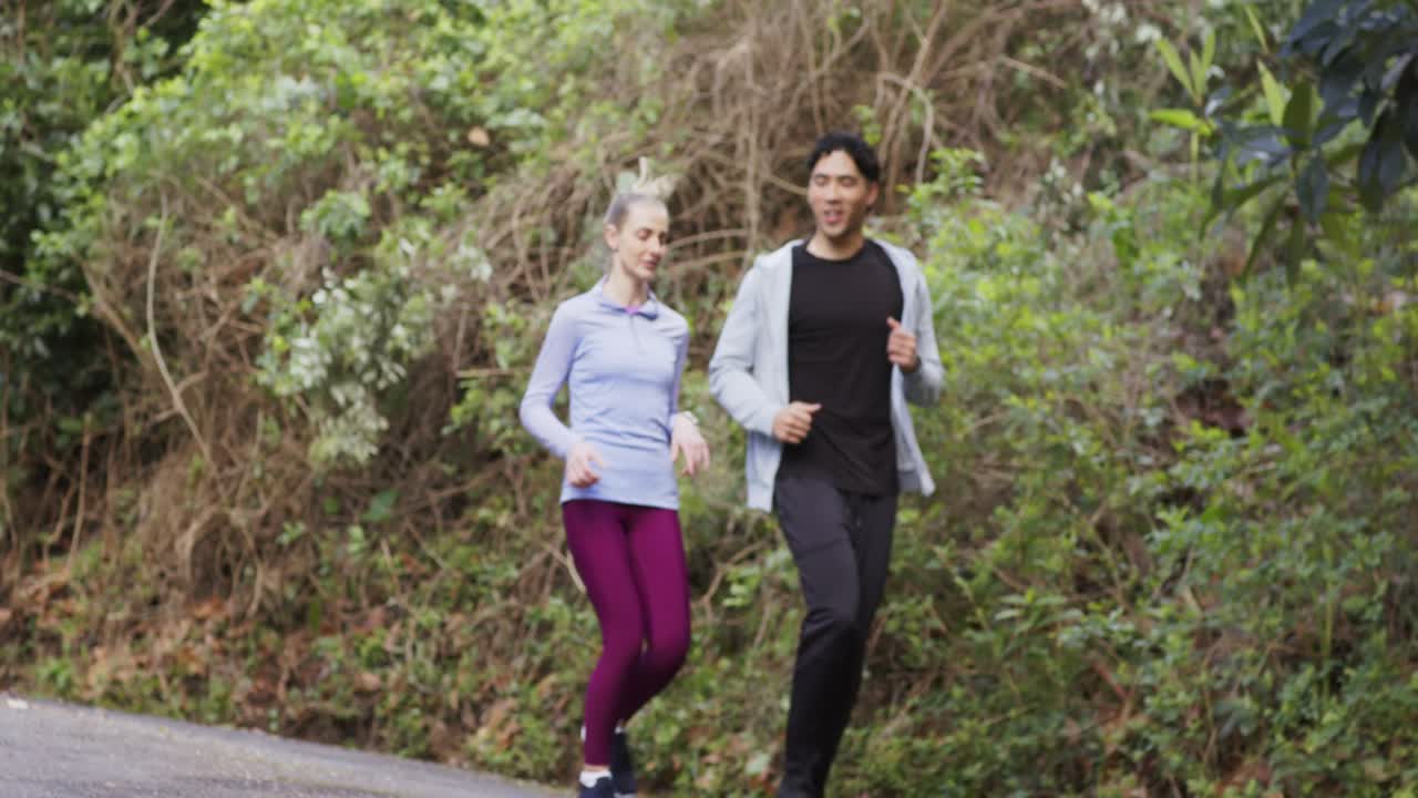 Cielo coperto che motiva una coppia di diverse etnie a fare jogging in abbigliamento sportivo per mantenersi in forma su un sentiero forestale lastricato.
