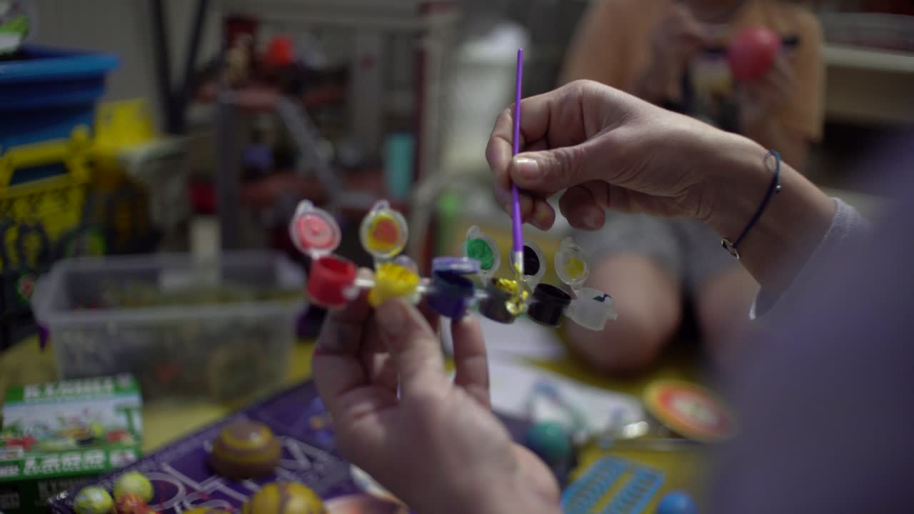 cerca de las manos de la mujer, mezclando colores de pintura con un pincel pequeño, su hijo pintando planeta de juguete en el fondo borroso en casa 4k