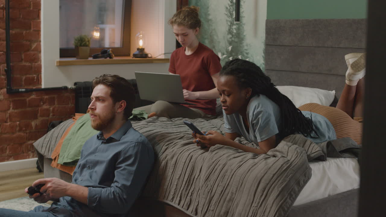 Three Rommates Using Smartphone, Playing Video Games And Working With Laptop On The Bed And Floor 1