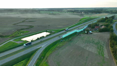 aerial view of a winding highway passing through green fields and farmlands, with a small pond and scattered rural houses in the background