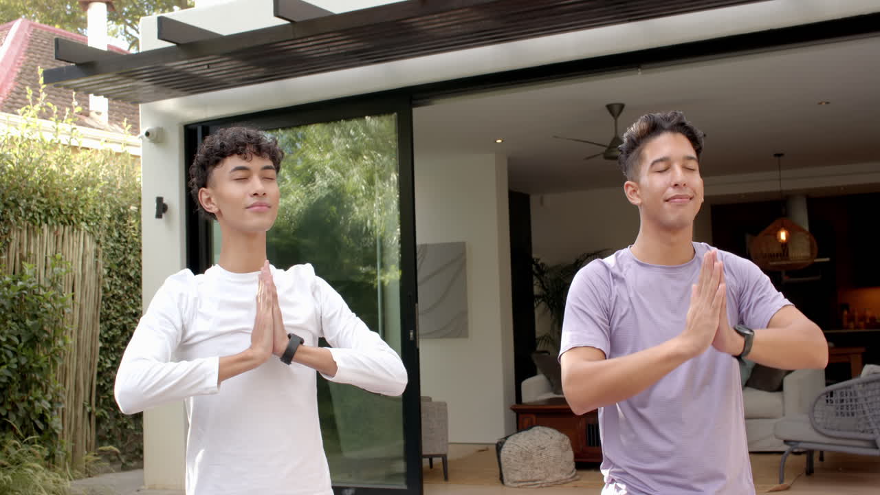 Practicing yoga, multiracial gay couple meditating with hands in prayer position outside home