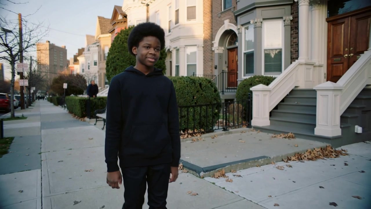 Walking teen wearing black hoodie and pants starting step on rowhouse sidewalk, passing iron fence