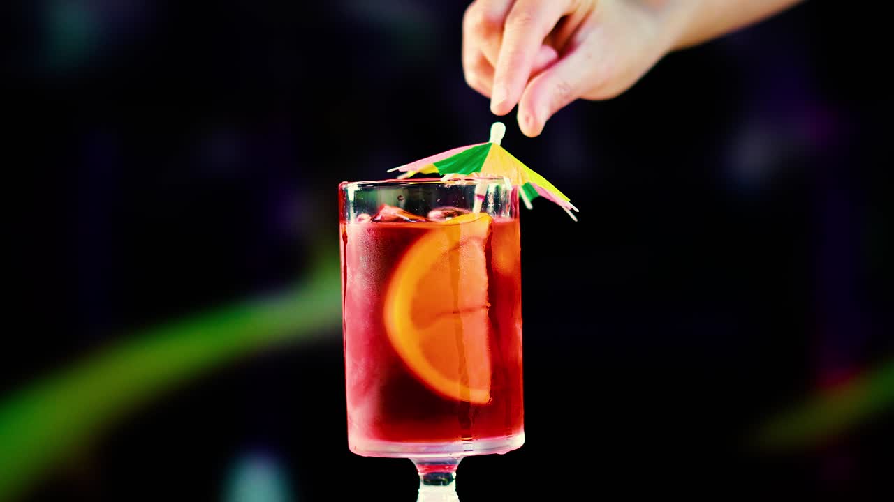 A Negroni cocktail is garnished with a colorful umbrella against a blurred background, highlighting vibrant colors and festive mood