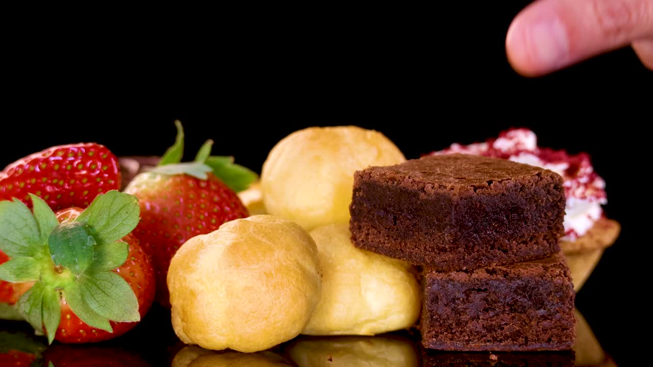 A hand selects a chocolate brownie from a vibrant dessert assortment including strawberries, cream puffs, and pastries, under bright studio lighting with a black background
