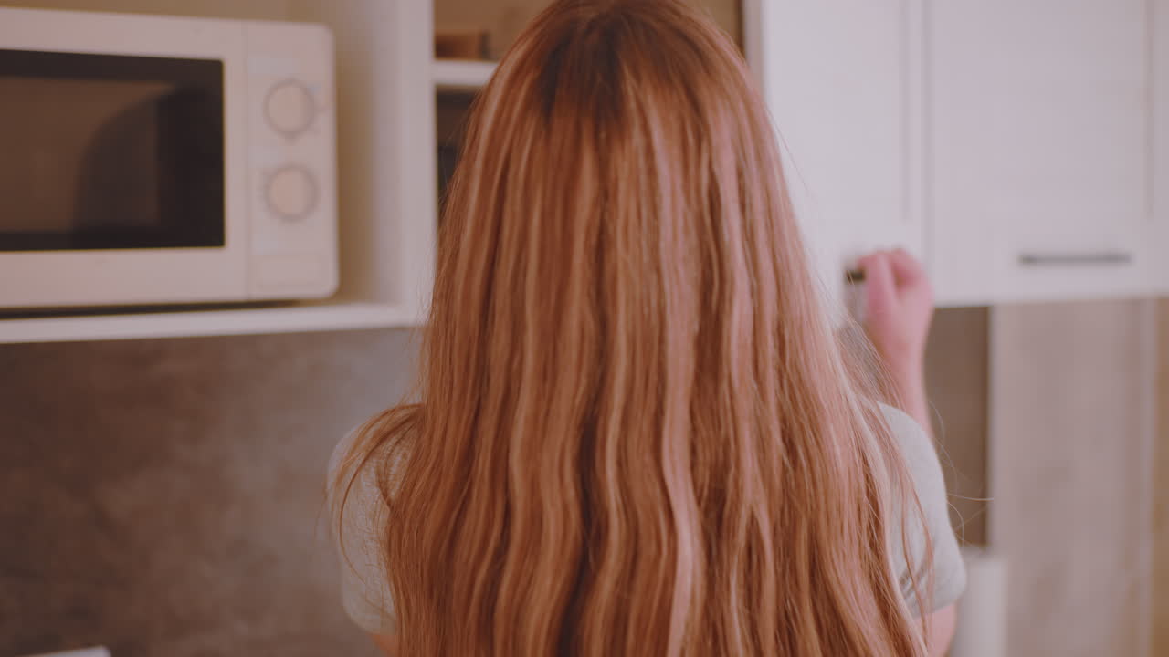 Natural light streams into kitchen as back view of woman in black short pants with flowing hair walks toward cabinet to collect item, creating serene casual household morning moment