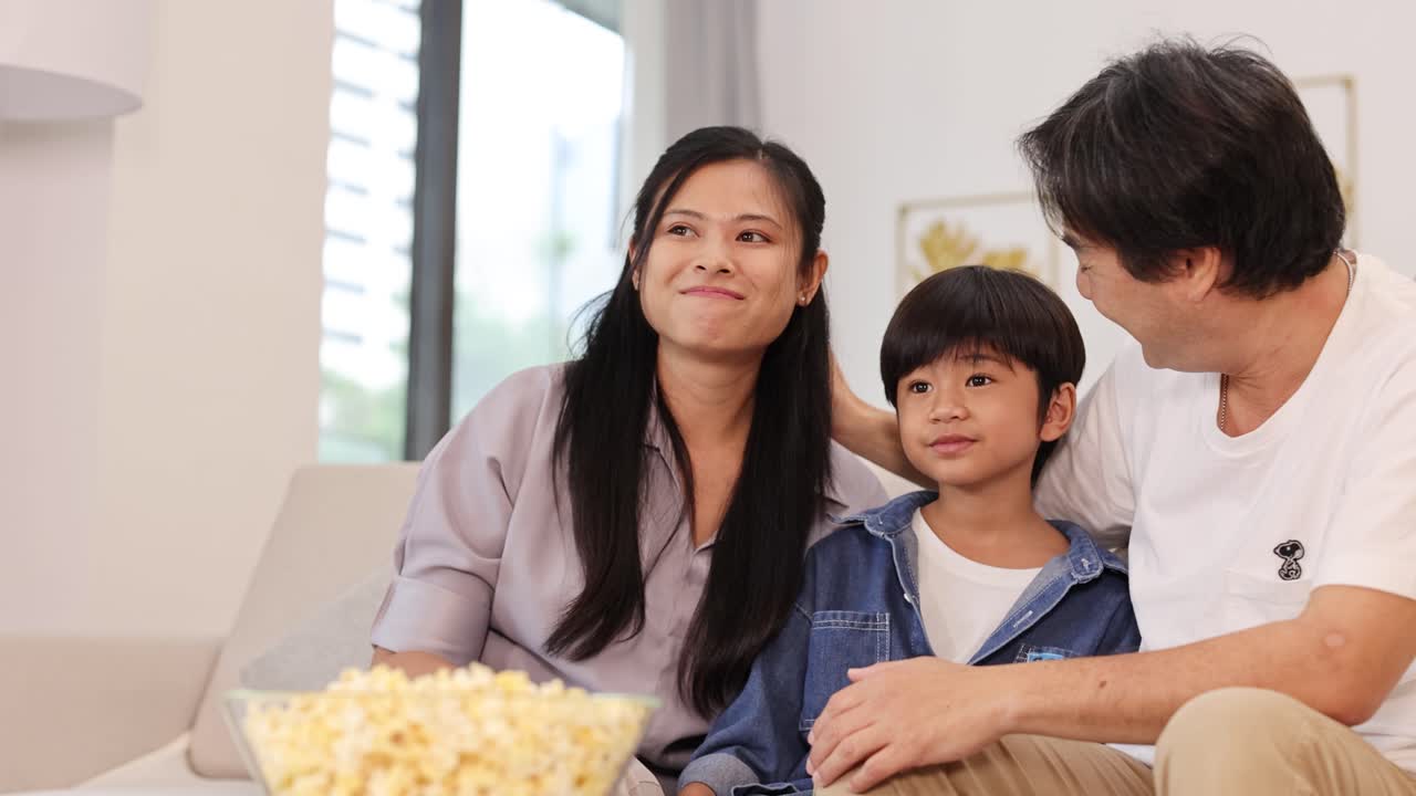 Happy Asian family bonding on couch, sharing popcorn in cozy, well-lit living room together