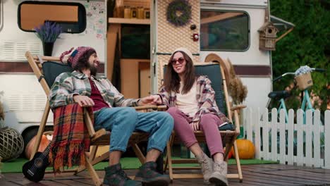 Happy-brunette-girl-in-round-glasses-and-a-white-hat-in-a-plaid-shirt-and-pink-pants-sits-on-a-chair-with-her-brunette-boyfriend-in-a-hat-near-a-trailer-during-their-picnic-at-a-camp-outside-the-city-in-the-summer