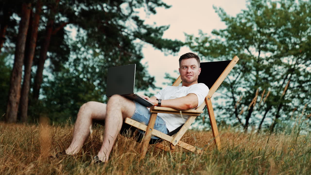 Positive smiling freelance male works in a comfortable chair. Remote work on the internet in the nature.