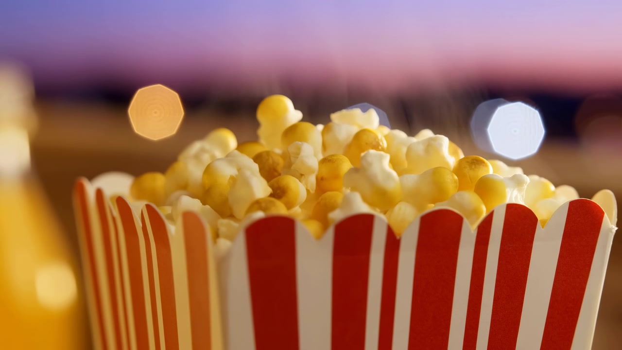 Una vista ravvicinata di un secchiello a strisce rosse e bianche pieno di popcorn in primo piano, sullo sfondo di un bokeh morbido e caldo con luci scintillanti al crepuscolo.