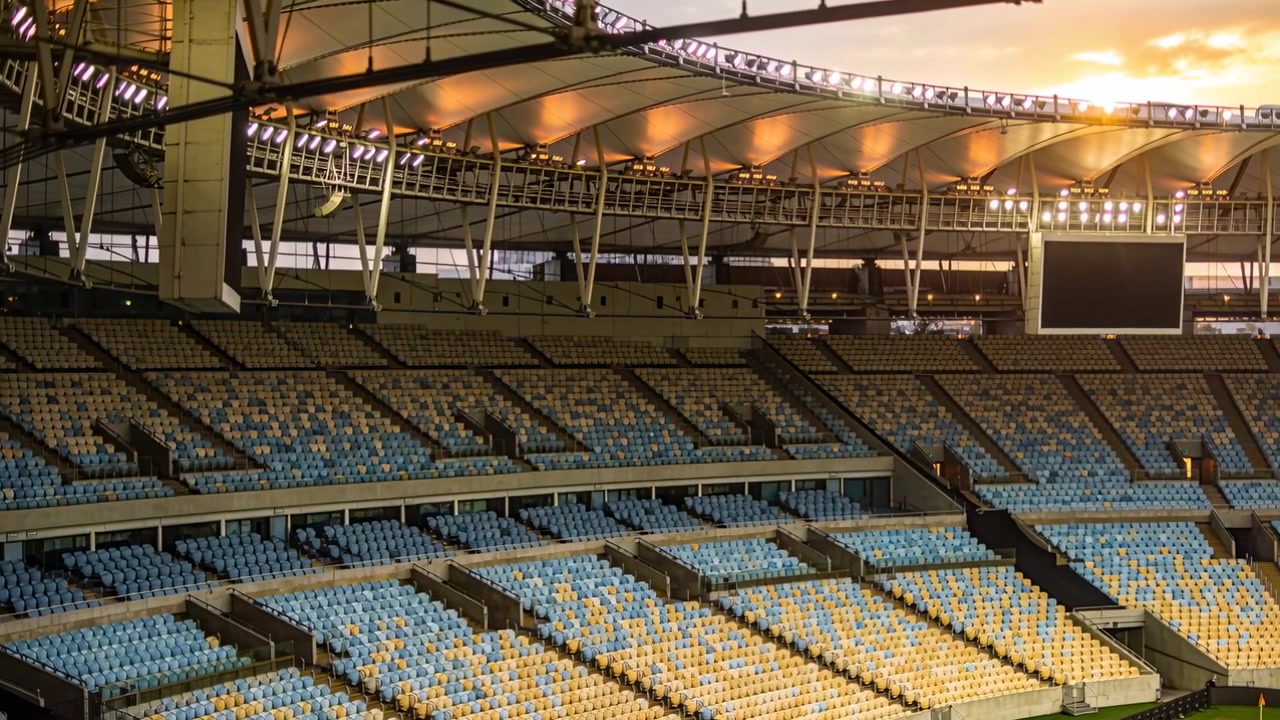 Shifting sunset light bathing empty blue yellow tiered stands in covered stadium, scoreboard unlit