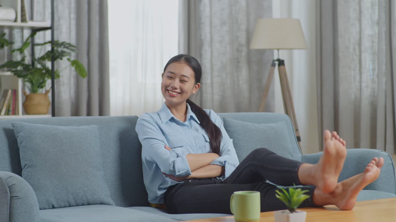 아시안 여성의 몸이 가득 차 있습니다. 나무 테이블 위에 발을 놓고 거실의 소파에 누워 있습니다.
