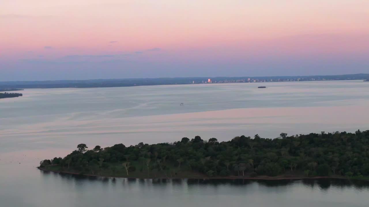 Wide drone shot capturing the river, an island, and the distant city lights under a vibrant pink and purple twilight sky