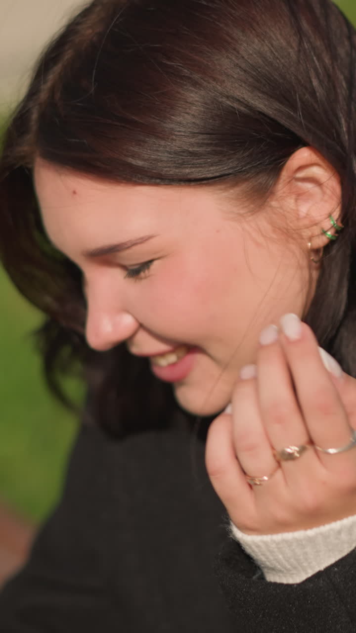 primer plano de una mujer con abrigo negro sonriendo mientras se ajusta el cabello en un parque público, pendientes visibles, dedo adornado con un anillo elegante, entorno al aire libre brillante con vegetación suave en el fondo