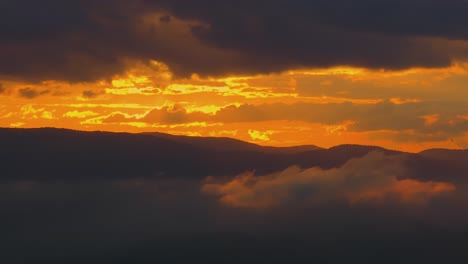 Aerial-close-up-shot-showing-an-orange-sunset-as-it-casts-a-warm-glow-between-a-cloudy-sky-and-and-distant-mountain-ranges,-high-contrast