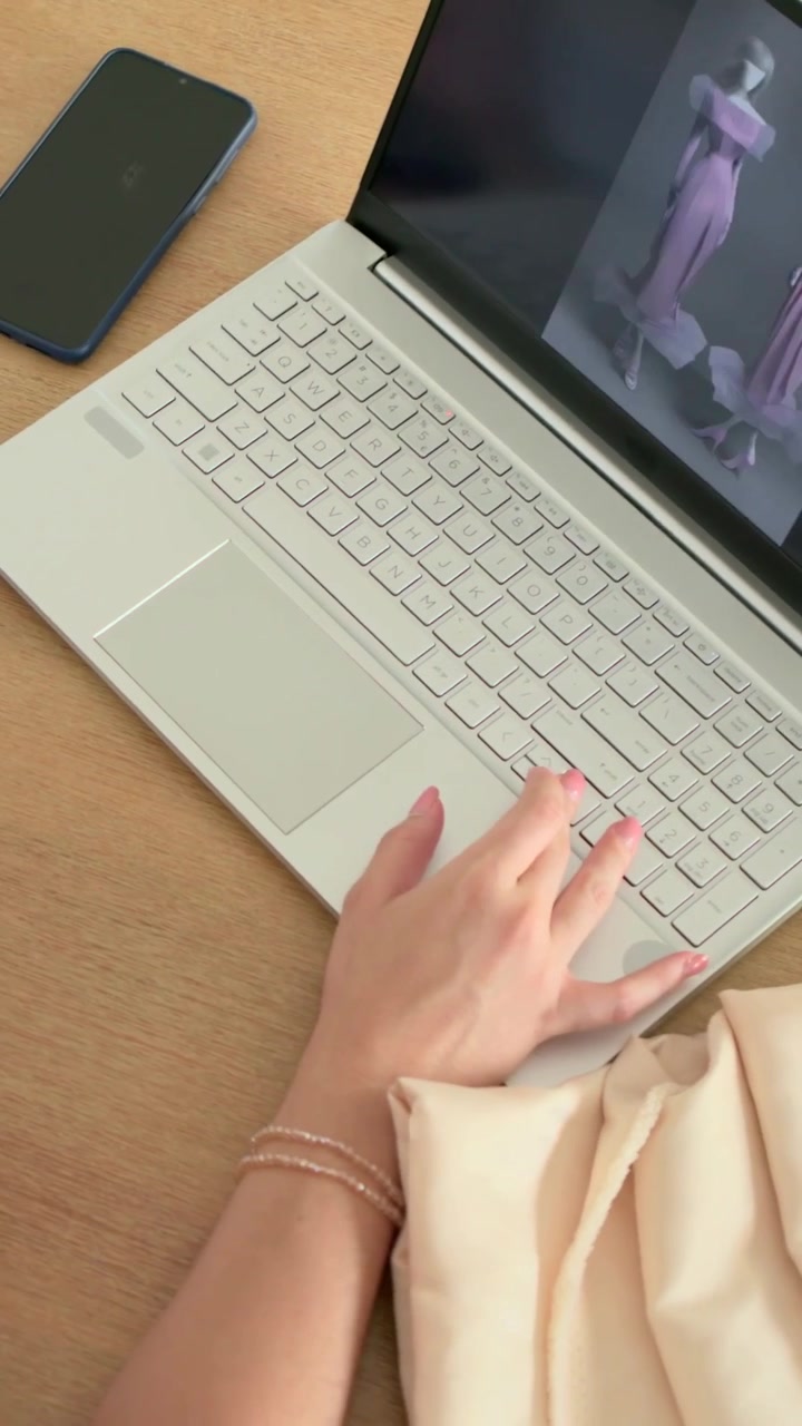 Video verticale: Donna con top color ruggine che si allunga verso il laptop sulla scrivania abbinando un tessuto rosa, prende appunti