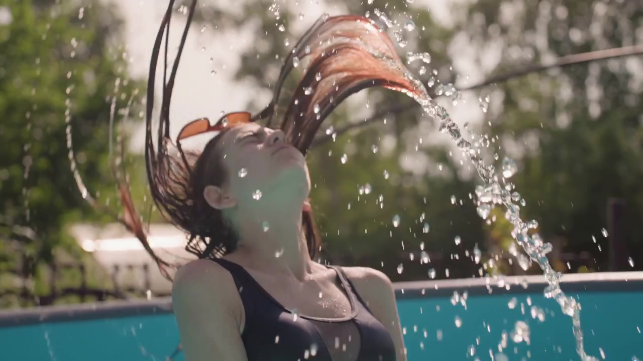Una mujer en traje de baño negro emerge del agua, se echa el pelo hacia atrás salpicando y bebe agua de la piscina. La escena se desarrolla al aire libre bajo la luz brillante del sol, con un entorno de piscina azul claro.