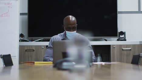 African-american-businessman-in-face-mask-using-laptop-in-office-conference-room