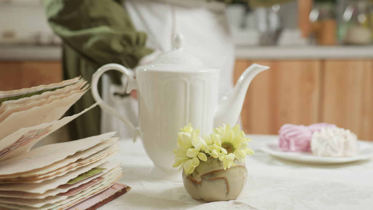 Close up of vintage open book on table beside small flower pot and ceramic jug as female cook places glass mug down gently before sitting