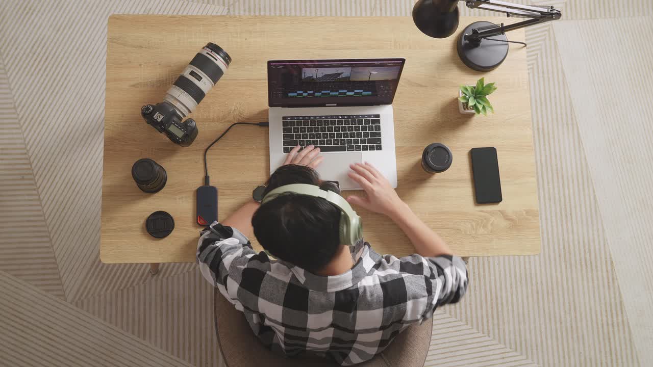 vista superior de un editor masculino asiático con auriculares que tiene dolor de espalda mientras está sentado en el espacio de trabajo usando una computadora portátil junto a la cámara editando el video en casa