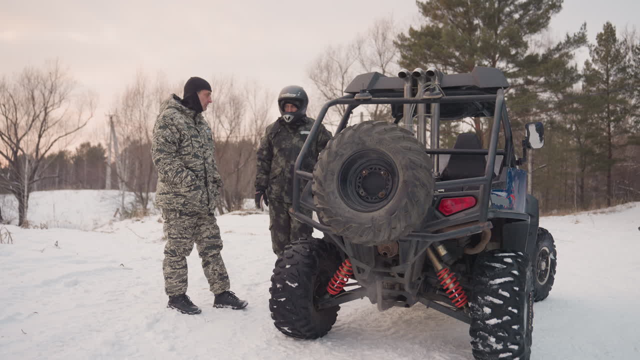Duo su ATV che conversa accanto a un UTV sulla neve in una radura fredda, passeggero con casco all'interno e pilota mimetico all'esterno, discussione del percorso e controlli di sicurezza con ruota di scorta visibile, bagliore del tramonto e attesa impaziente