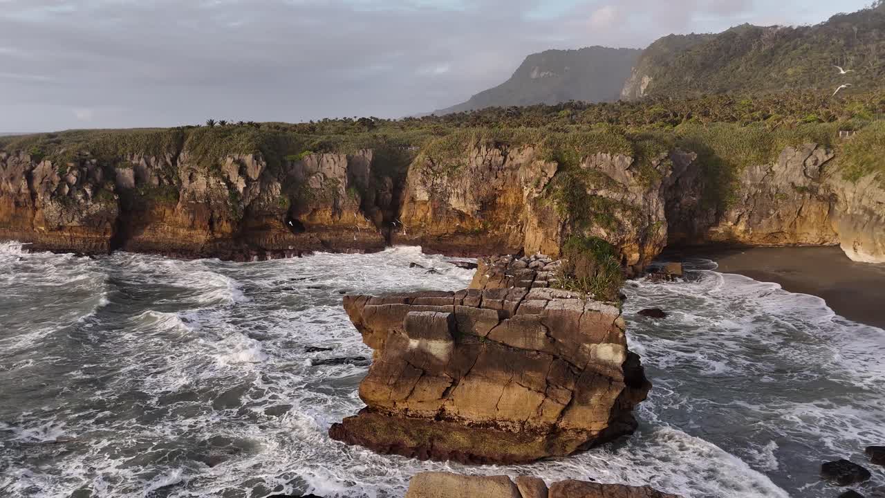 Faraglione marino e costa frastagliata con onde che si infrangono al tramonto vicino a Punakaiki, Nuova Zelanda. Ripresa aerea all'indietro.