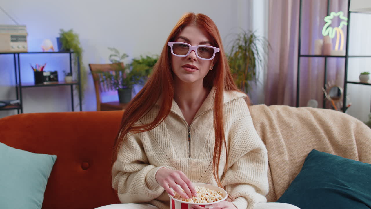 Woman sitting on couch eating popcorn and watching interesting tv 3d film sport game online at home