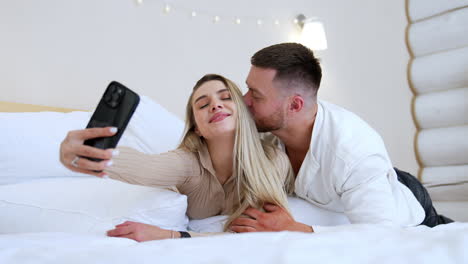 caucasian woman with long blonde hair lies on the bed near her man. girl is taking selfie as the man kisses her on the cheek.