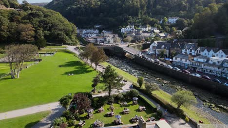 Aerial-Footage-of-East-Lyn-river-and-park-Lymouth-Devon-UK-cars-crossing-road-bridge-people-sitting-outside-pub-for-lunch