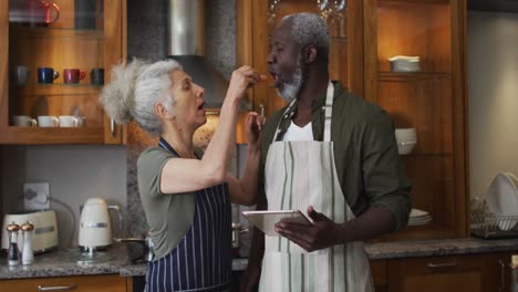 caucasian senior woman wearing apron feeding chopped vegetable to her husband in the kitchen at home