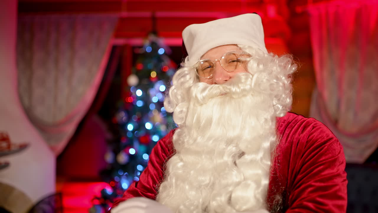 Santa Claus smiling on camera indoors. Portrait of a real Santa with white beard, in glasses and red costume standing in the living room on Christmas tree background.