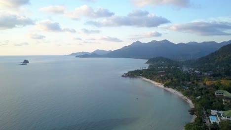 Una-Toma-Aérea-Durante-Una-Puesta-De-Sol-Desde-Un-Dron-Sobre-La-Costa-De-Una-Isla,-En-La-Distancia-Hay-Montañas-Visuales