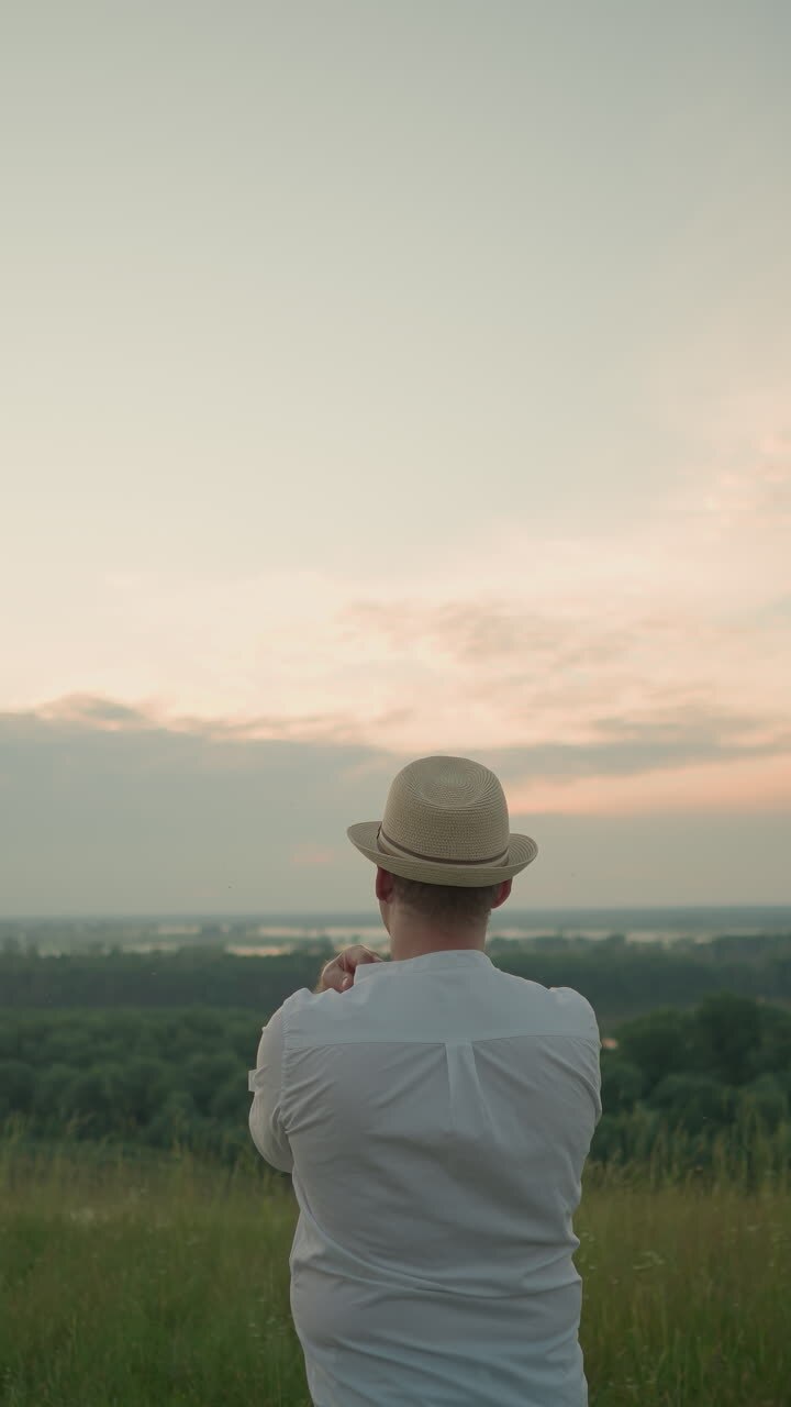 A thoughtful man wearing a white shirt, hat, and jeans stands in a grassy field by a tranquil lake at sunset, gazing into the distance. The warm sunset light create a peaceful atmosphere