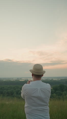 un hombre pensativo con una camisa blanca, sombrero y vaqueros está de pie en un campo cubierto de hierba junto a un lago tranquilo al atardecer, mirando hacia la distancia. la cálida luz del atardecer crea una atmósfera pacífica