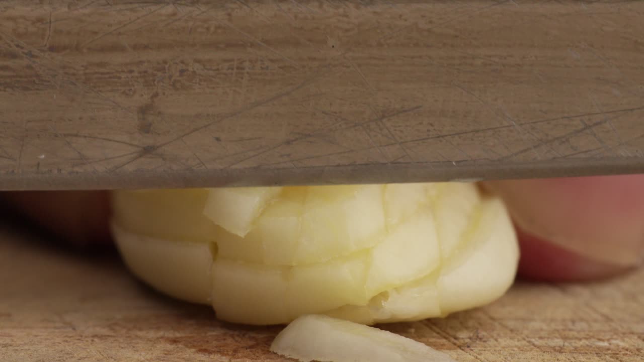 Macro of mincing garlic slice cloves on dark wood surface in dramatic lighting with high detail and shadows as knife cuts downward