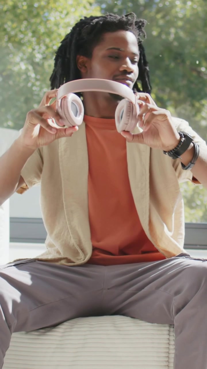 Vertical video: Lifting pink headphones, man in rust adjusting at window removing to look outside