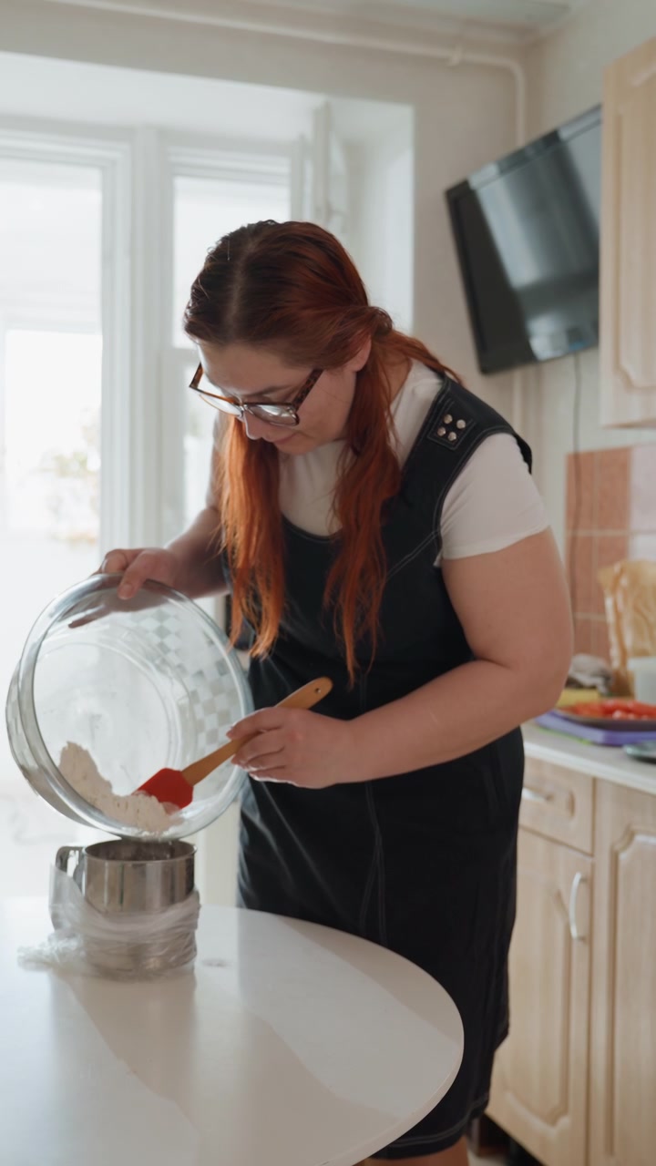 Mulher inclina-se sobre a ilha da cozinha, inclinando uma tigela de vidro enquanto usa uma espátula para raspar massa espessa para uma panela, mostrando um movimento cuidadoso de despejar, mãos firmes, luz natural brilhante, culinária caseira.