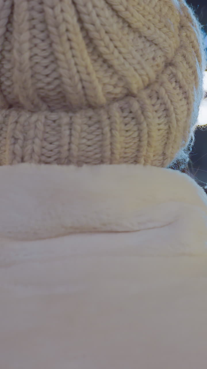 Close-up of woman in warm winter beanie standing still while people skate and move around under glowing decorative lights, with illuminated Christmas tree and blurred city background