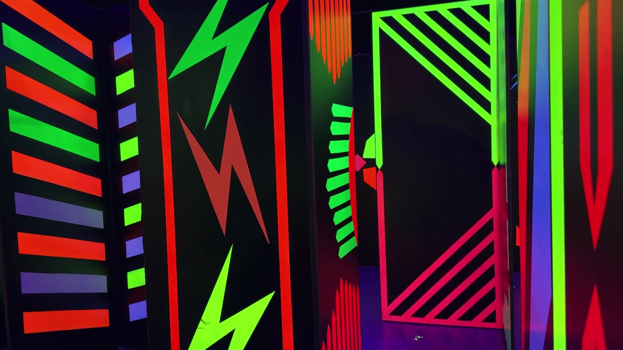 Labirinto interno buio con pannelli a parete neri dipinti con forme geometriche e fulmini luminosi e vivaci di colore verde, rosso e arancione, illuminati da luce nera UV in un'arena di laser tag