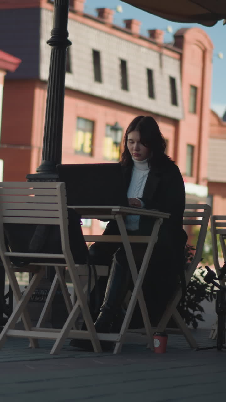Tech-savvy woman working on laptop outdoors at cafeteria, surrounded by wooden chairs, plants, and tripod, seated at table with coffee cup, paved walkway in background