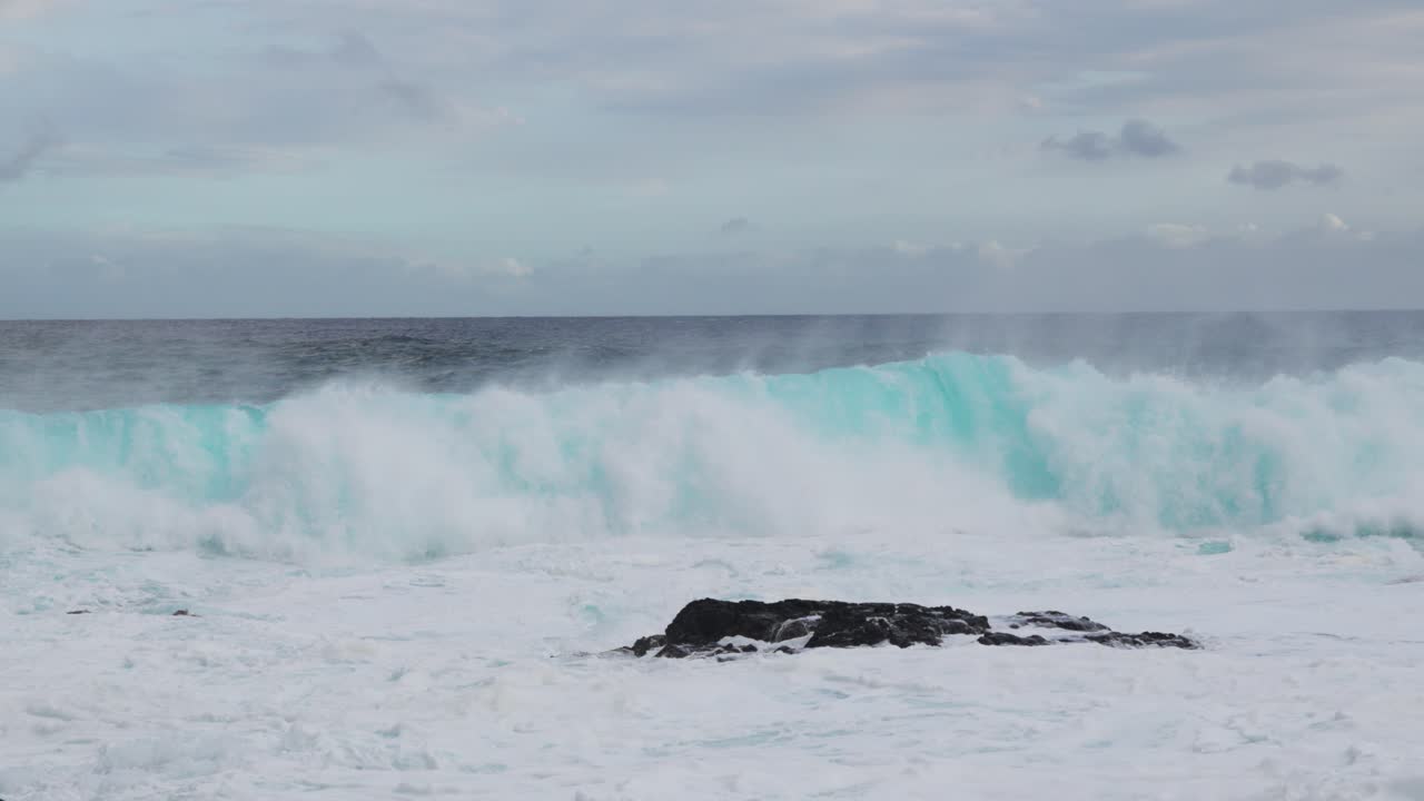 A teal set lifts and folds just offshore as wind tears spindrift from the crest, while thick whitewater blankets dark shore rocks beneath a mottled, cloud-streaked sky