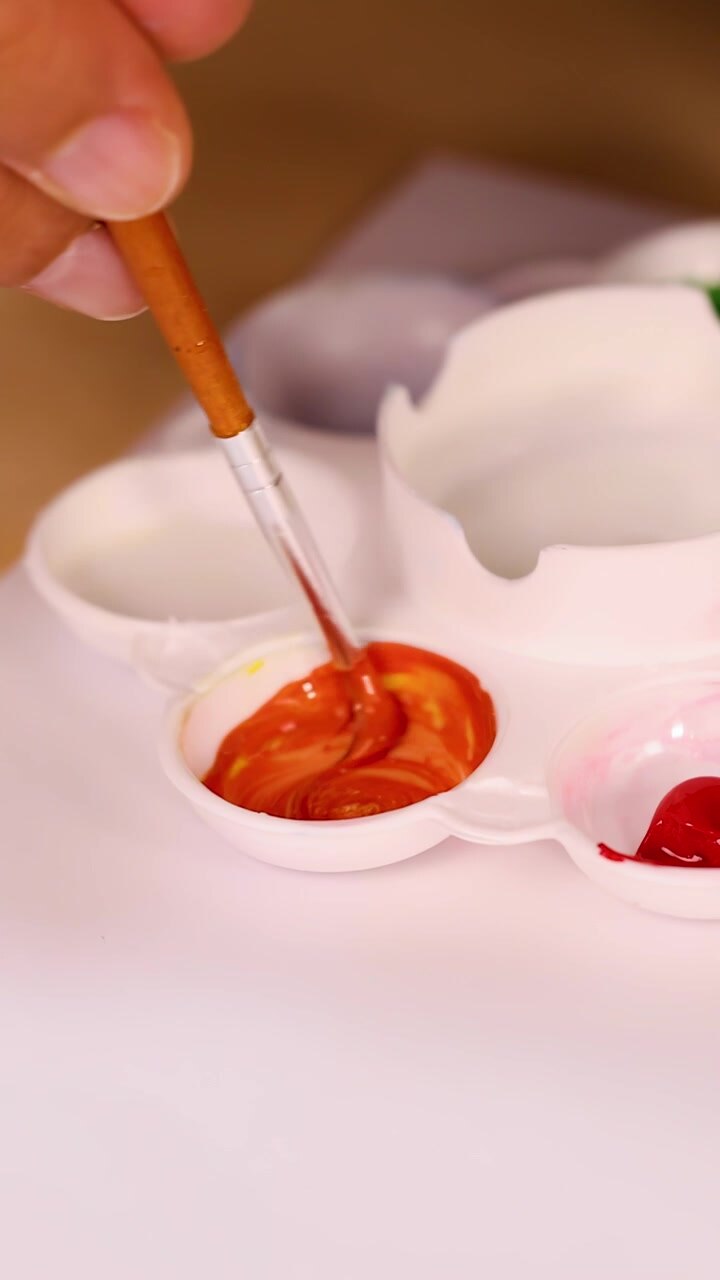 Artist’s hand mixes red and yellow watercolor paints in palette under warm, soft lighting
