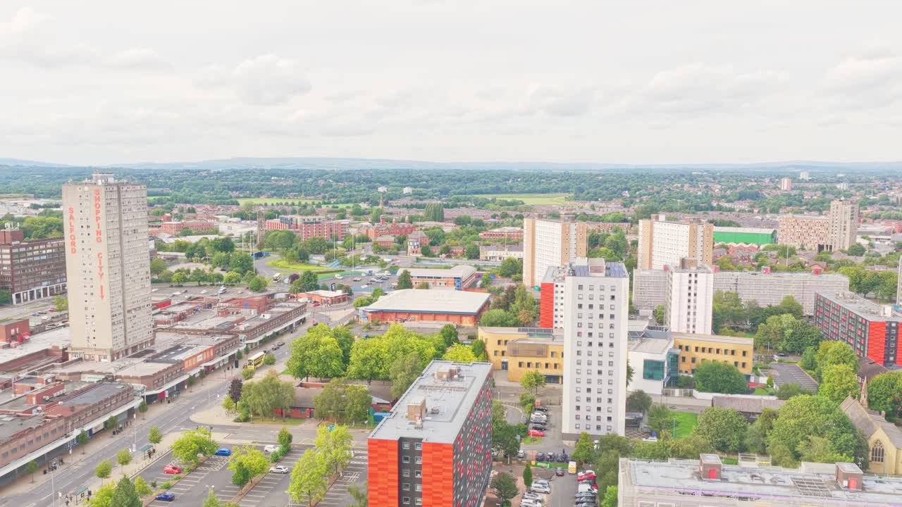 Diverse torri residenziali a molti piani a Salford si ergono tra edifici a uso misto, strade alberate e viste aperte verso colline lontane, catturate in una ripresa aerea ascendente con drone.