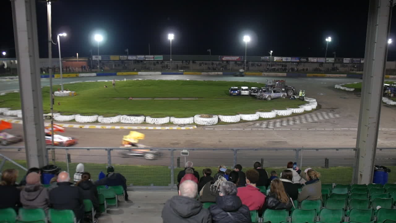 Spectators watching as stock cars race around the track under bright floodlights, with cars blurring past in the foreground. The open-air stadium provides a lively view of the high-speed night event