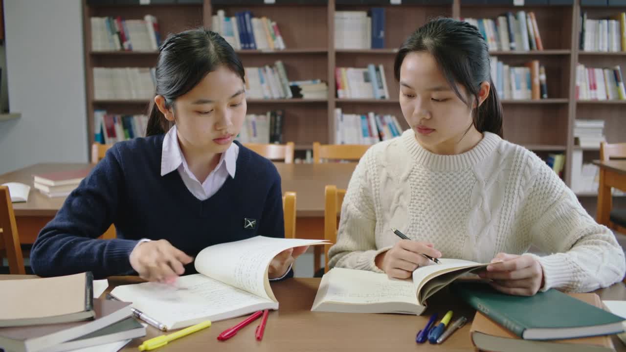 図書館で、左の紺色のセーターを着た人がワークブックを指しながらページをめくり、右のクリーム色のセーターを着た人がそれを見ている。