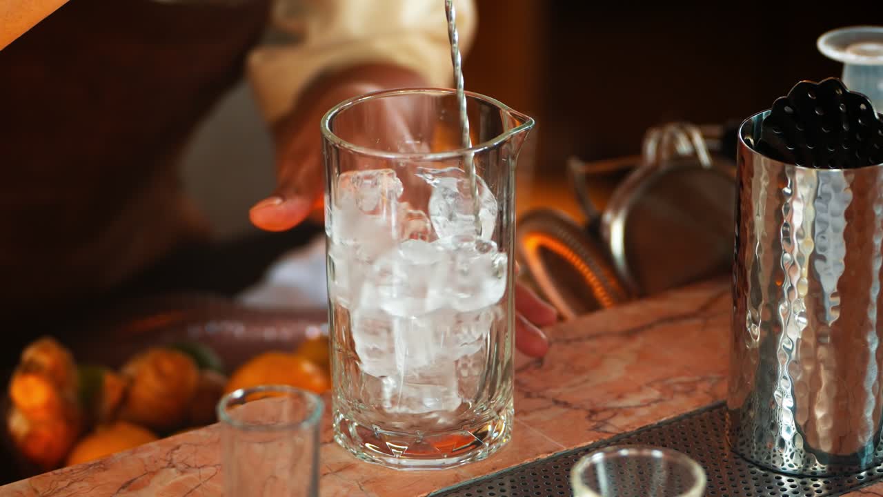 Skilled bartender hands using a long bar spoon to stir ice cubes, chilling a mixing glass in preparation for crafting a classic cocktail at an elegant bar during lesson - close-up